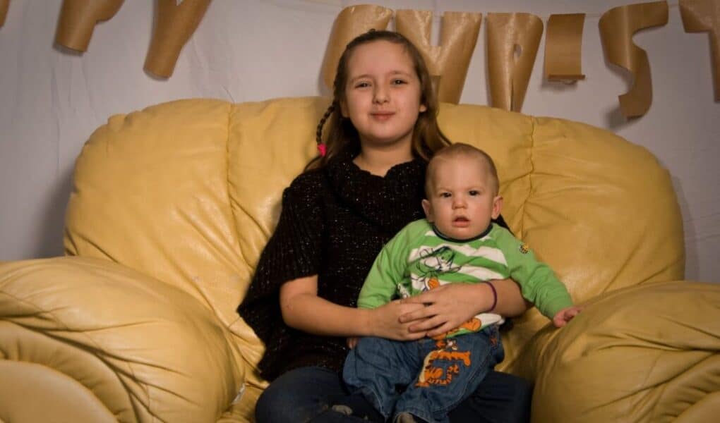 A girl with long brown hair sits on a large yellow armchair, smiling, with a younger baby boy in a green shirt on her lap. Behind them hangs a banner with partially visible letters, likely spelling “Happy Christmas”.
