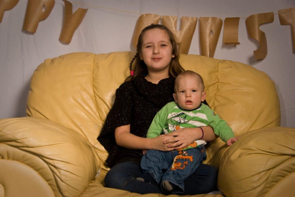 A young girl with brown hair sits on a large yellow armchair, holding a baby boy in her lap. Behind them is a white backdrop with brown paper letters, partially reading “CHRIST.” Both children look towards the camera, the girl smiling gently.