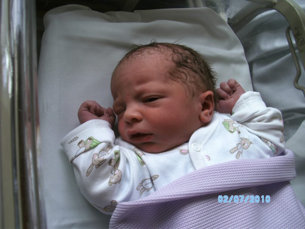 A newborn baby with dark hair lies on a white blanket in a hospital cot, wearing a white babygrow with bunny prints and wrapped in a light purple blanket. The baby’s eyes are partially open. The photo is dated 07/02/2010.