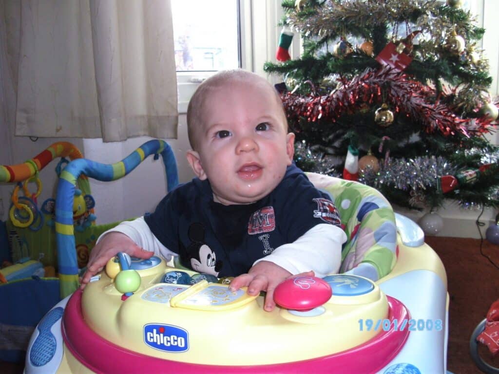 A baby sits in a colourful Chicco activity walker, smiling and looking up. Behind the baby is a decorated Christmas tree with baubles and tinsel. A window with net curtains lets in daylight. The date "19/01/2008" is visible in cyan text at the bottom right.