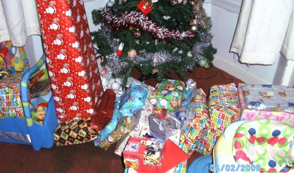 A Christmas tree decorated with tinsel and baubles stands in the corner, surrounded by many wrapped presents in colourful paper. A pink and green baby seat and a toy box are nearby. The date stamp on the photo reads "04/02/2008".
