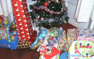 A Christmas tree decorated with tinsel and baubles stands in the corner, surrounded by many wrapped presents in colourful paper. A pink and green baby seat and a toy box are nearby. The date stamp on the photo reads "04/02/2008".
