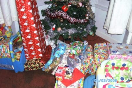 A Christmas tree decorated with tinsel and baubles stands in the corner, surrounded by many wrapped presents in colourful paper. A pink and green baby seat and a toy box are nearby. The date stamp on the photo reads "04/02/2008".