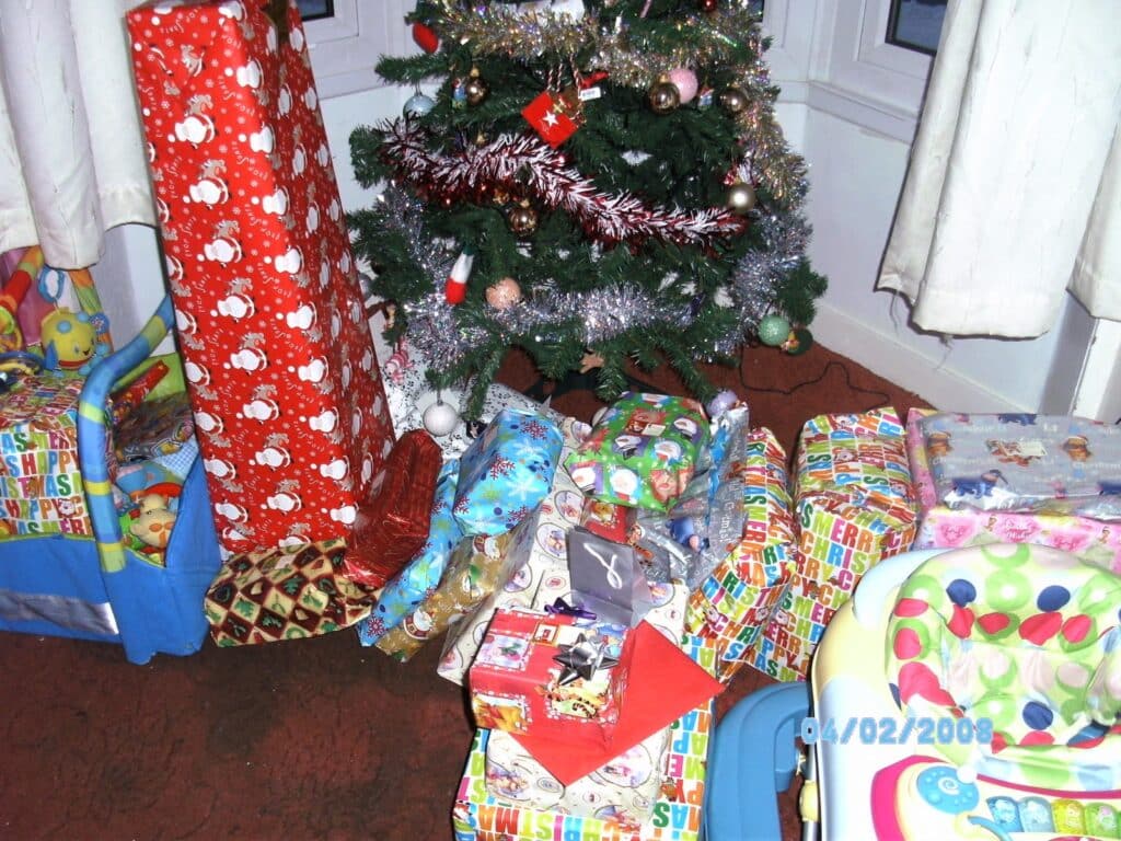 A Christmas tree decorated with baubles and tinsel stands by a window. Piles of festively wrapped presents surround the tree, along with a toy pushchair and a colourful baby seat. White curtains frame the scene. The date stamp reads 04/02/2008.