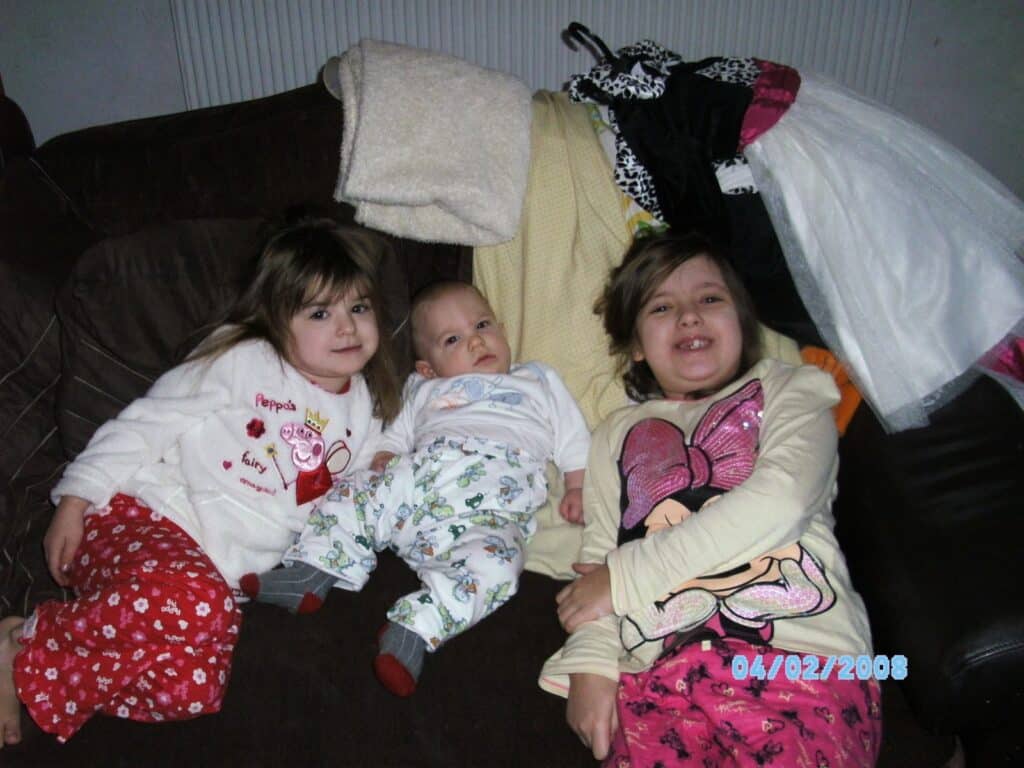 Three children relax on a dark settee: two girls in patterned pyjamas and a baby in white and blue pyjamas. The girl on the left wears red Peppa Pig trousers; the girl on the right has a Minnie Mouse top. Clothes and blankets are piled in the background. Date: 04/02/2008.