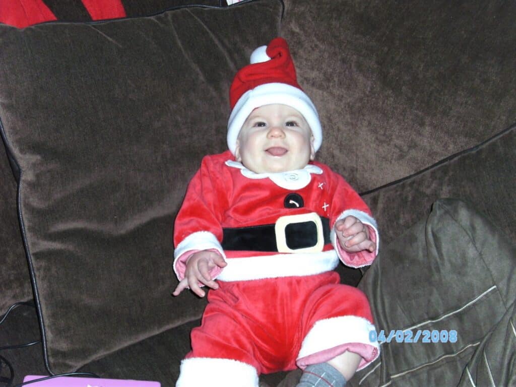 A smiling baby wearing a red and white Father Christmas outfit and hat sits on a brown sofa. The baby’s hands are open, and the outfit features a black belt and white trim. The date “04/02/2008” is displayed in blue text on the bottom right.