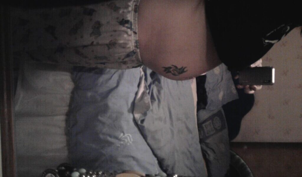 A person stands sideways in front of a mirror, showing their bare midriff with a tattoo on the right side of their lower abdomen. They are wearing light-coloured pyjama shorts with a print. The reflection reveals a bed with blue bedding in a dimly lit room.