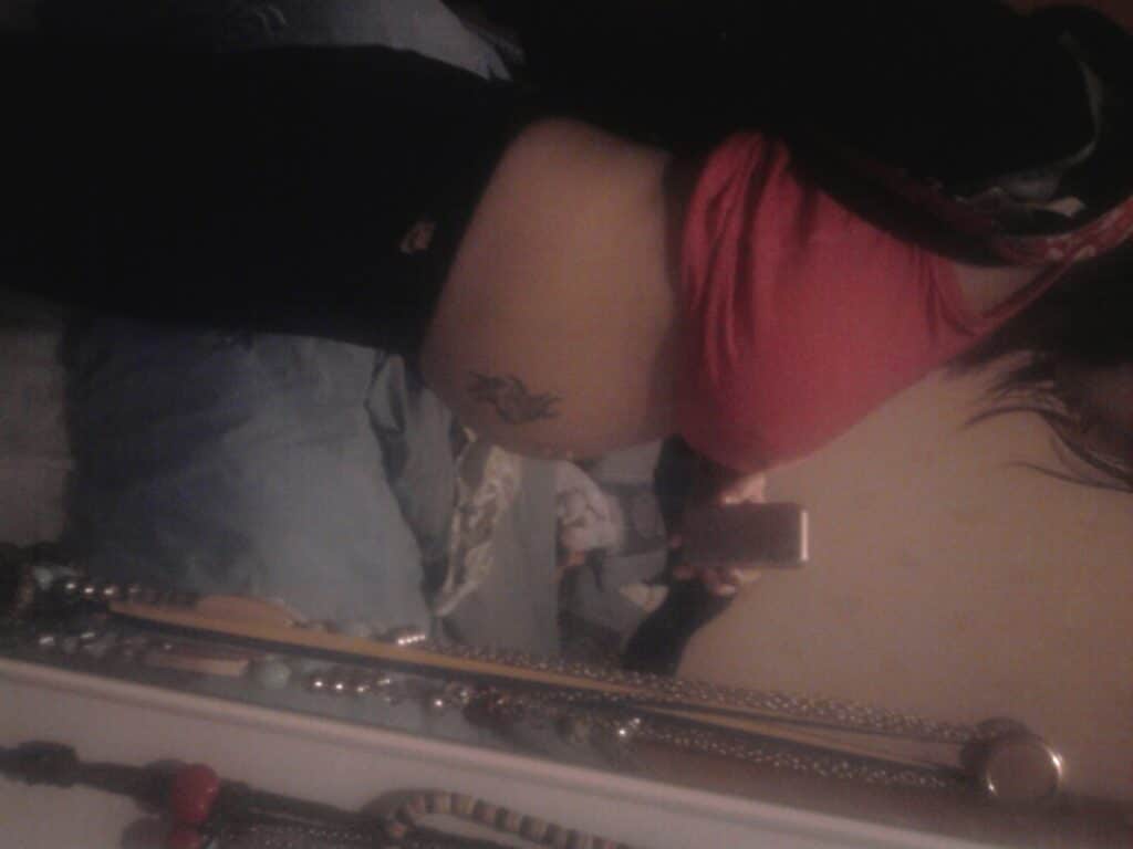 A person in a red top and black trousers takes a mirror selfie, showing a tattoo on their side. The focus is on their midsection, possibly highlighting a pregnant belly. Jewellery and accessories hang on the mirror’s edge, and a bed is visible in the background.