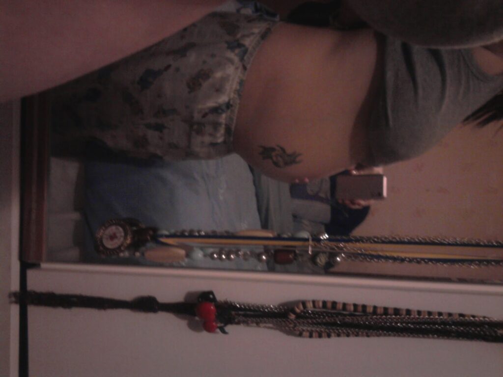 A person takes a wing mirror selfie, showing their midriff with a floral tattoo on their side. They wear a grey crop top and patterned shorts. The mirror reflects a chest of drawers with jewellery and personal items on it. The lighting is dim.