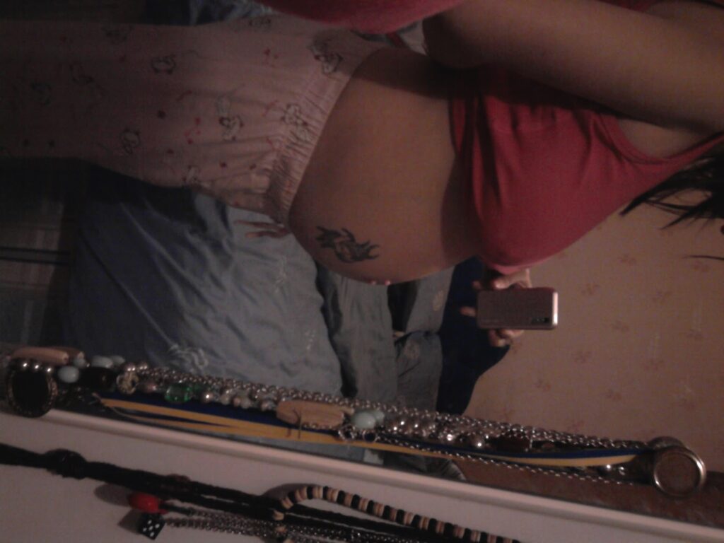 A person wearing pink pyjama trousers and a red top takes a mirror selfie, showing a side view of their partially exposed stomach with a tattoo. The background includes a made bed with blue sheets and a white chest of drawers with jewellery.
