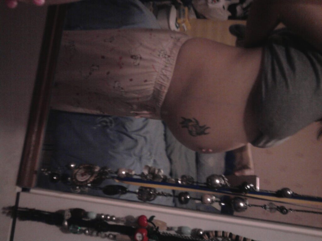 A person is standing sideways in front of a mirror, wearing light-coloured pyjama trousers and a grey top. Their midsection and tattoo on their hip are visible. The room in the background includes a bed and scattered items. Necklaces hang from the mirror's edge.