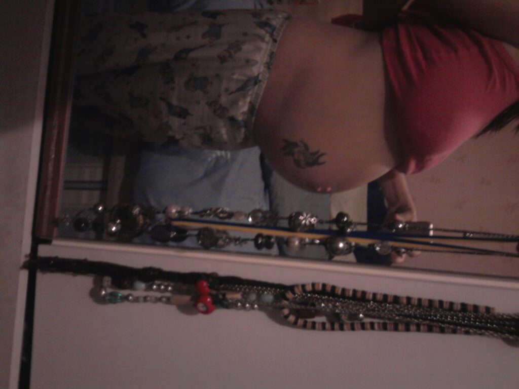A person stands sideways in front of a mirror, showing a pregnant belly with a tattoo on the side. They are wearing pyjama bottoms and a cropped pink top. Necklaces and other jewellery hang on the mirror's frame in the foreground. The image is dimly lit.