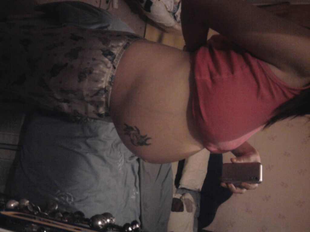 A person in patterned pyjama trousers and a pink top is taking a side mirror selfie, showing their bare pregnant belly with a dragon tattoo. The photo is indoors, near a bed, and the person is holding a phone to capture the image.