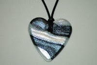 A heart-shaped pendant with black, white, and silver swirled stripes hangs from a black cord against a plain, light-coloured background. The pendant has a shiny, reflective surface.