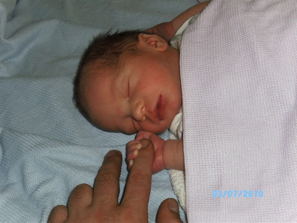 A newborn baby with dark hair sleeps on a light blue blanket, wrapped in a pale pink cloth. The baby gently grips an adult’s finger with its tiny hand. The date “03/07/2010” is visible in blue text at the bottom right.