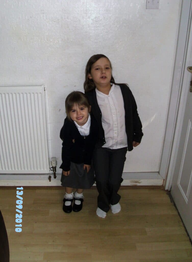 Two young girls in school uniforms stand indoors on a wooden floor by a white wall and radiator. The younger girl, smiling, wears a dark cardigan, grey skirt, and black shoes, while the older girl, serious, wears grey trousers and white socks. Date: 13/09/2010.