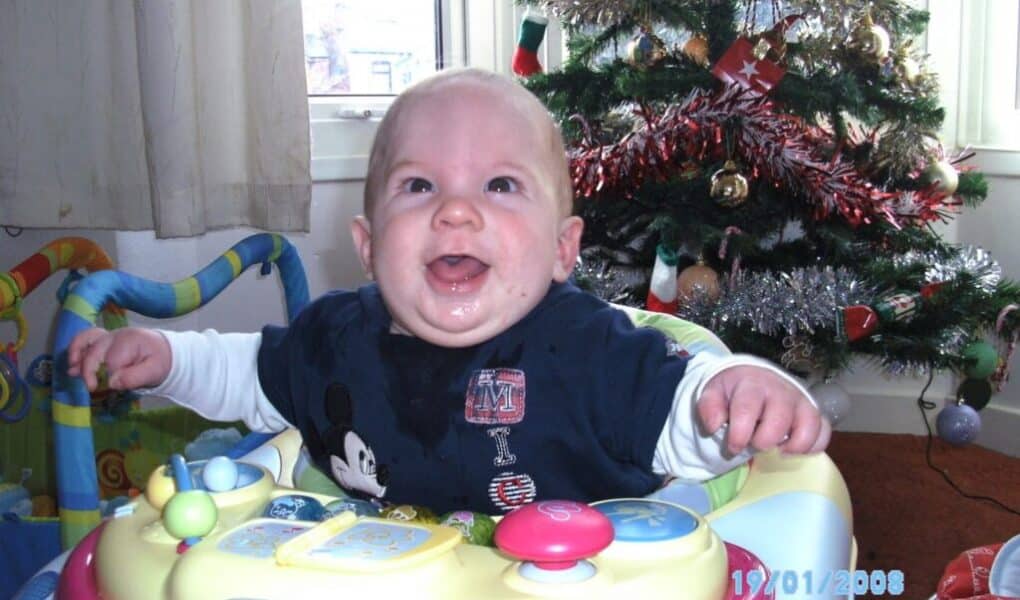 A smiling baby sits in a Chicco walker in front of a decorated Christmas tree with tinsel and baubles. The baby wears a navy blue Mickey Mouse shirt and white sleeves. Sunlight shines through a window, and colourful toys are visible in the background.