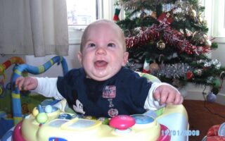 A smiling baby sits in a Chicco walker in front of a decorated Christmas tree with tinsel and baubles. The baby wears a navy blue Mickey Mouse shirt and white sleeves. Sunlight shines through a window, and colourful toys are visible in the background.