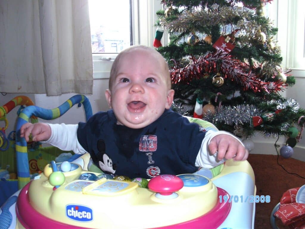 A smiling baby in a blue shirt sits in a yellow and green Chicco activity walker. Behind the baby is a decorated Christmas tree with tinsel and baubles, and a window with curtains letting daylight in. The date "19/01/2008" is displayed on the photo.