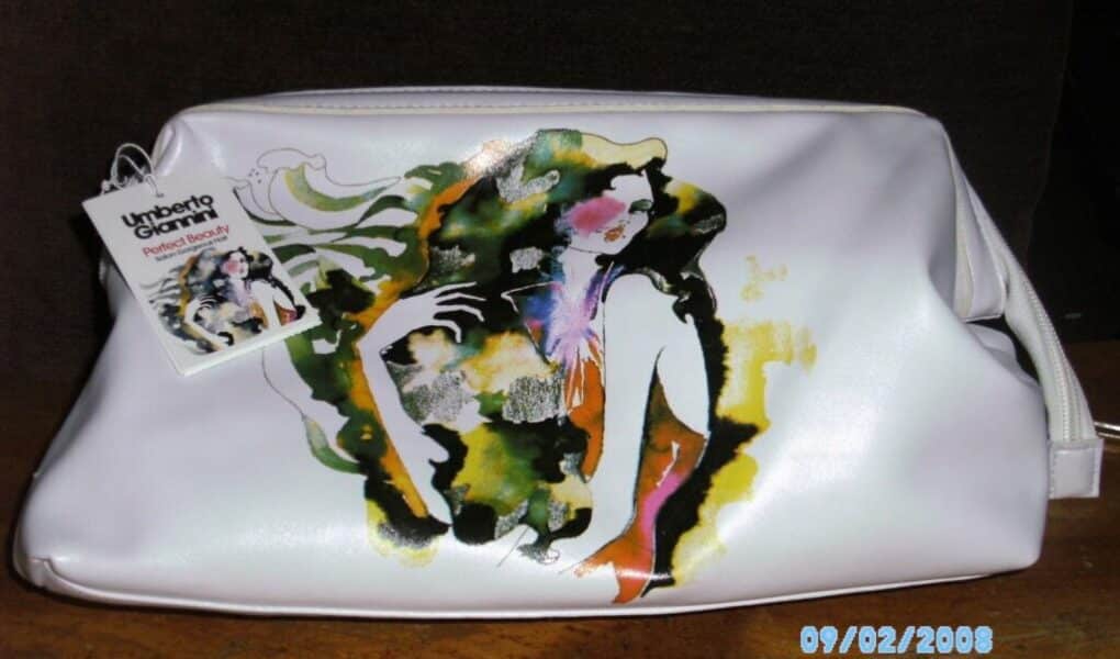 A white cosmetic bag with an abstract, colourful illustration of a woman on the front. A tag with the Umberto Giannini Beauty logo and product info is attached to the bag. The photo is dated 09/02/2008 and has a dark brown background.