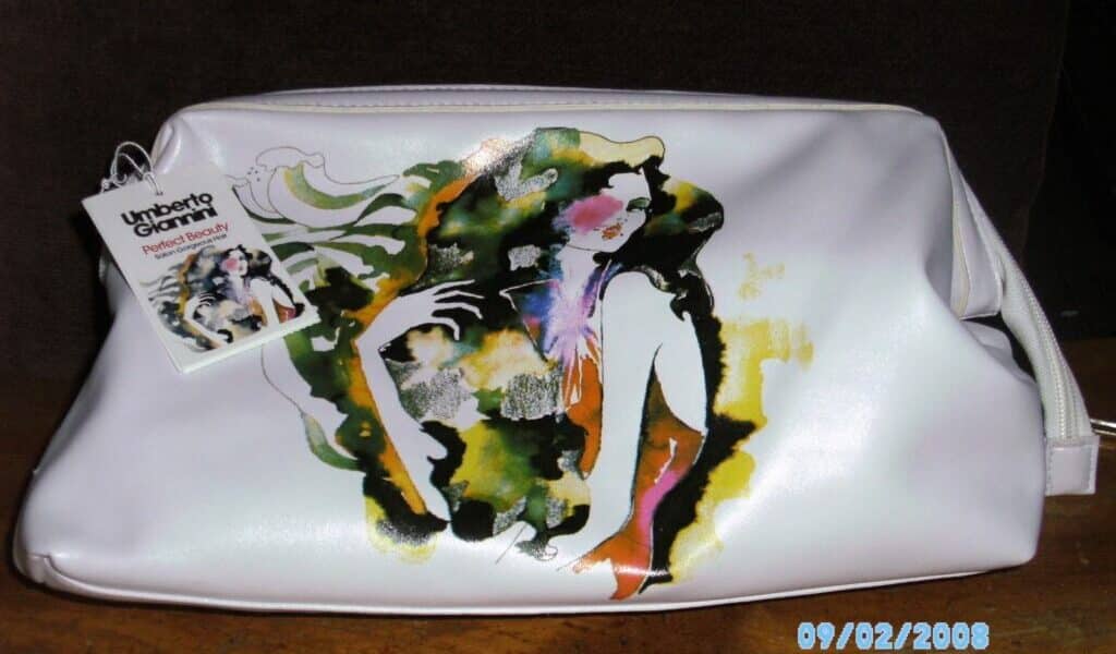 A white cosmetic bag with an abstract, colourful illustration of a woman on the front. A tag with the Umberto Giannini Beauty logo and product info is attached to the bag. The photo is dated 09/02/2008 and has a dark brown background.