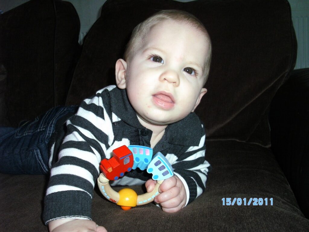 A baby with short fair hair, wearing a black-and-white striped jumper and jeans, lies on a brown sofa, holding a colourful toy train ring. The baby looks upwards with a neutral expression. The date "15/01/2011" is displayed in the bottom right corner.