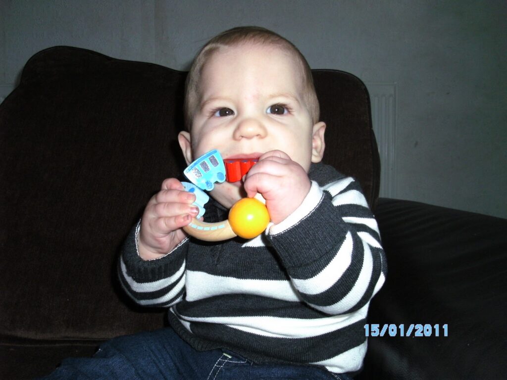 A baby wearing a black and white striped jumper sits on a dark sofa, holding a colourful toy with an orange ball and blue shapes. The baby looks at the camera. The date "15/01/2011" appears in blue text at the bottom right.