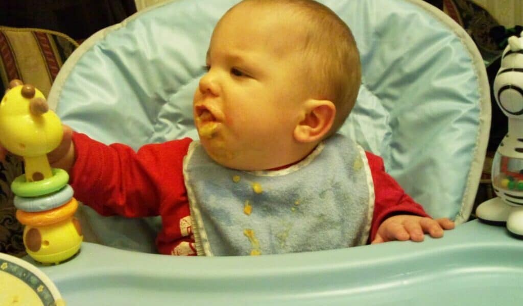 A baby wearing a blue bib with food stains sits in a high chair, looking to the left and holding a yellow toy with colourful rings. The baby has food on their mouth and cheek. A plate is partially visible in the bottom left, and a zebra toy is on the right.