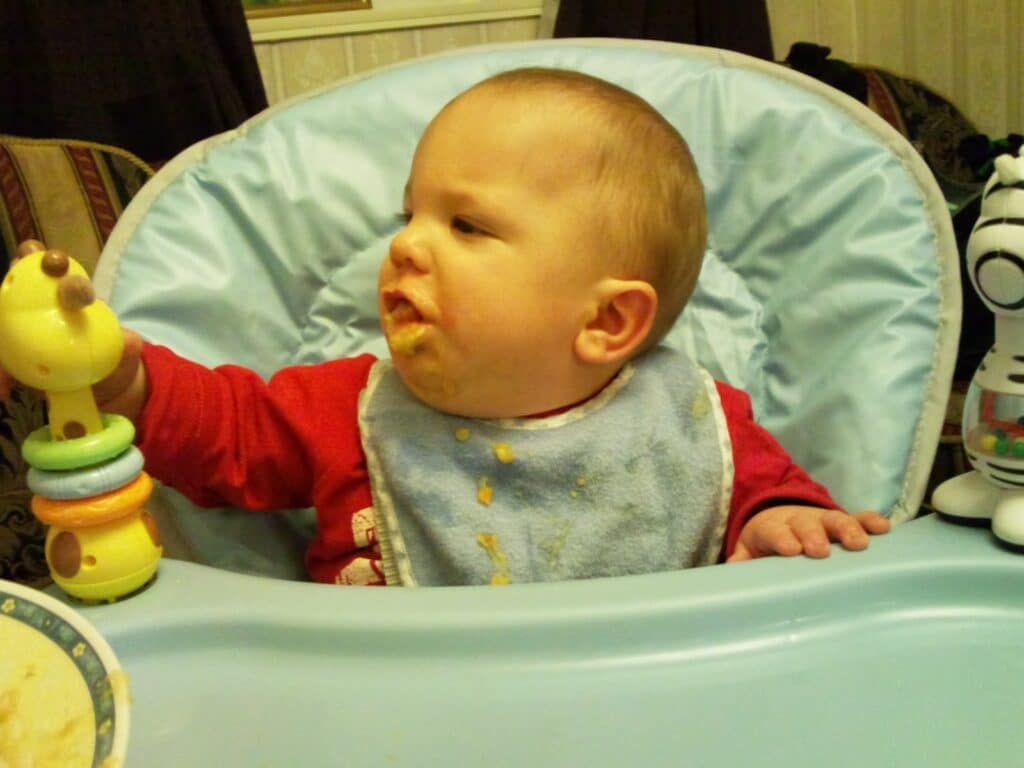 A baby wearing a red shirt and a blue bib with food stains sits in a highchair, holding a colourful toy in one hand and looking at it. There is baby food on their face and on the bib. A zebra-striped toy is on the tray to the right.