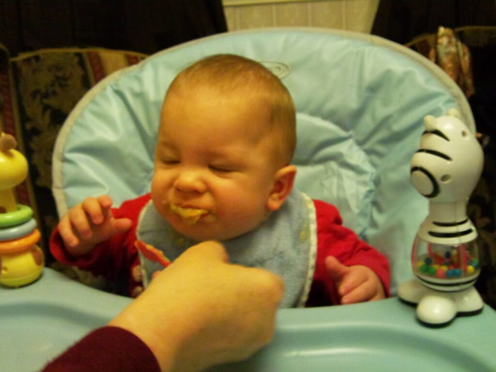 A baby with closed eyes and a messy face sits in a highchair, wearing a blue bib. An adult hand is feeding the baby, who appears to dislike the food. Colourful toys are attached to the highchair tray.