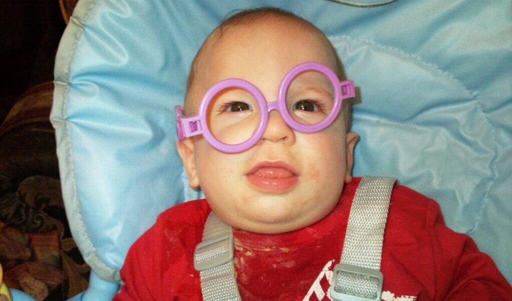 A baby wearing oversized, round, purple toy glasses sits in a light blue Graco highchair. The baby is dressed in a red shirt with white print and has some food stains on it. The baby looks forward with a slightly open mouth.