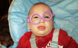 A baby wearing oversized, round, purple toy glasses sits in a light blue Graco highchair. The baby is dressed in a red shirt with white print and has some food stains on it. The baby looks forward with a slightly open mouth.