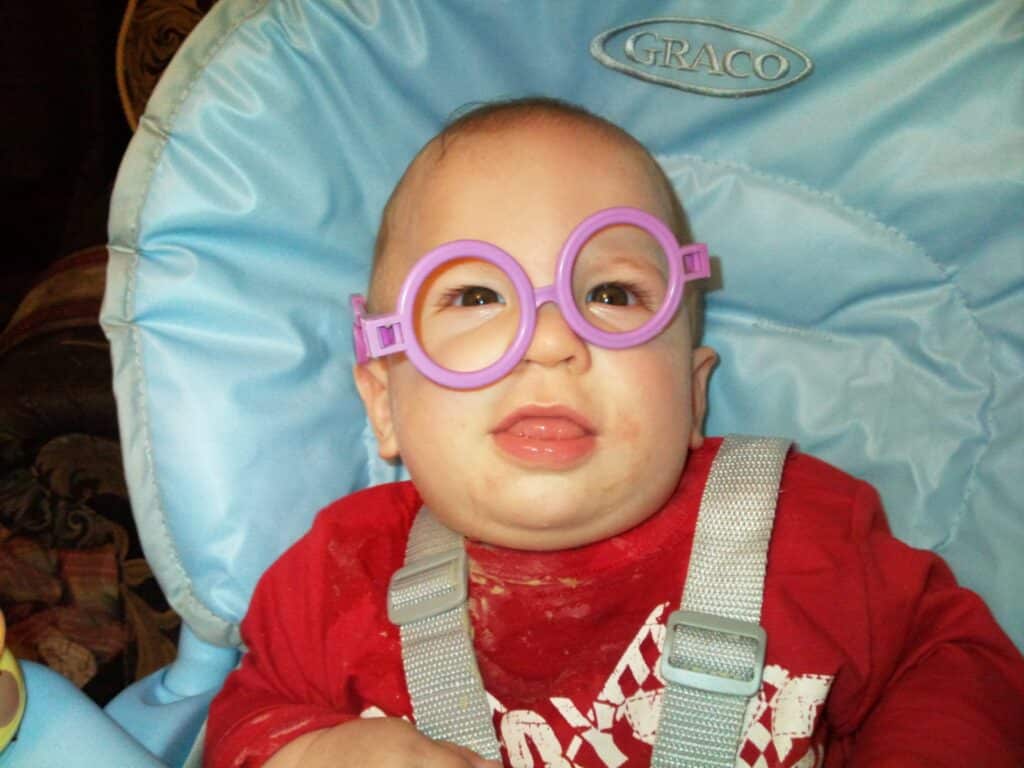 A baby sits in a light blue Graco highchair, wearing a red shirt with a white print and playful oversized purple round glasses. The baby’s mouth is slightly open, and there are food stains on the shirt.