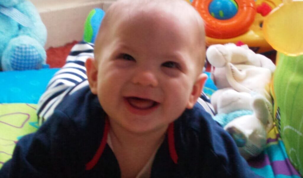 A smiling baby with light skin and short hair lies on a colourful play mat, surrounded by toys including a plush blue bear, a white soft toy, and a toy with buttons and a steering wheel in the background. The baby wears a dark top with red trim.