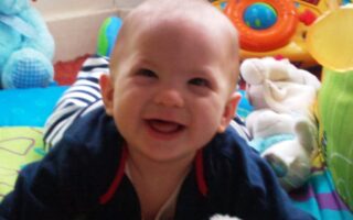 A smiling baby with light skin and short hair lies on a colourful play mat, surrounded by toys including a plush blue bear, a white soft toy, and a toy with buttons and a steering wheel in the background. The baby wears a dark top with red trim.
