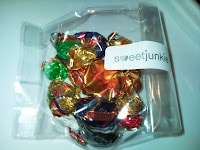A small, clear plastic box contains assorted colourful wrapped sweets. The sweets are mainly gold, red, green, and blue. A white label on the box reads "sweetjunkie." The box rests on a white surface.