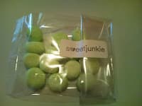A clear plastic bag filled with small, round, green sweets sits on a pale surface. The bag has a white label with the word “sweetjunkie” printed on it in lowercase letters. The sweets appear smooth and glossy.