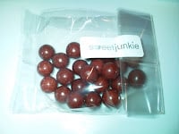 A clear plastic bag containing several round, shiny, brown sweets. The bag has a white label with the partially visible text "sweetjunkie." The background is a plain, light surface.