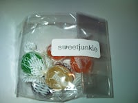 A clear plastic bag containing colourful round sweets—green, orange, yellow, and purple—wrapped in white netting. The bag has a white label with "sweetjunkie" printed in lowercase black letters on the front.