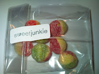A clear plastic bag contains six round, sugar-coated sweets, each half red and half yellow-green. A white label with "sweetjunkie" in black and green text is attached to the bag. The background is a plain white surface.