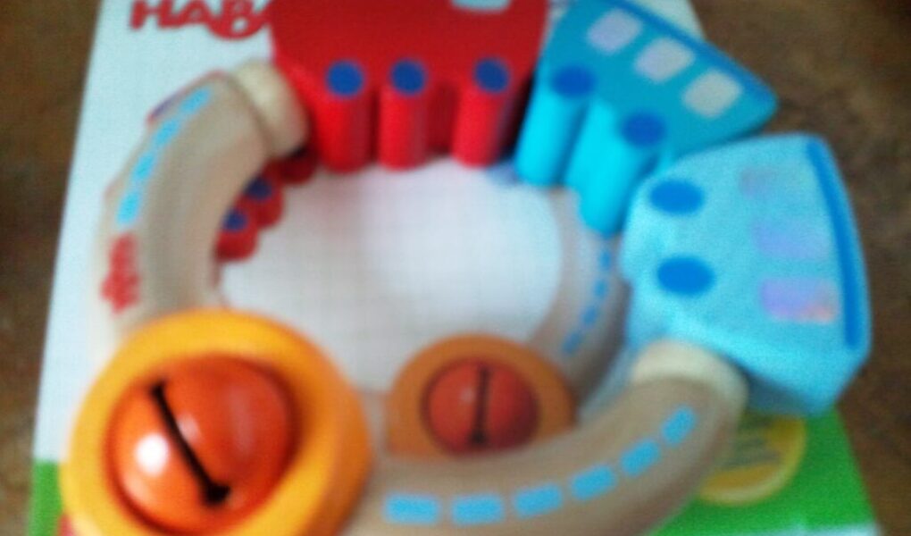 A colourful, circular wooden toy train set with red and blue train engines and two tan curved tracks. The toy features orange knobs with black arrows and is placed on top of a HABA-branded box. The image is slightly blurry.