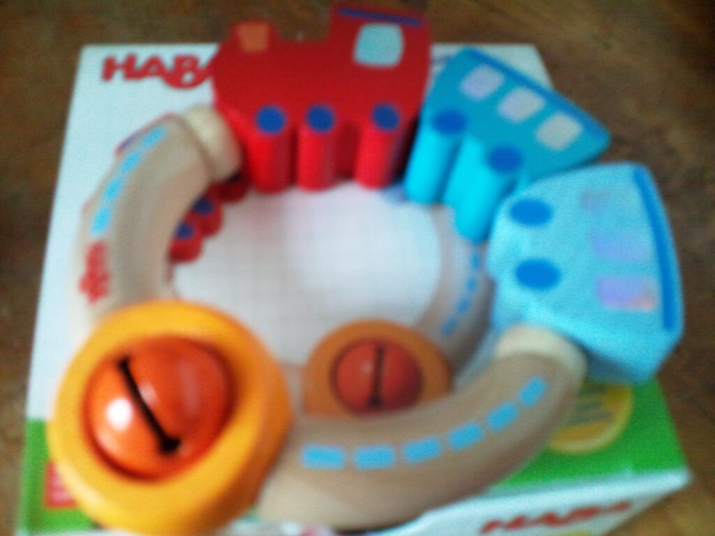 A colourful toy train set forms a ring with red and blue train carriages connected by beige tracks. Two orange circular pieces with black arrows resemble bells or clocks. The background shows a partially visible box with the HABA logo.