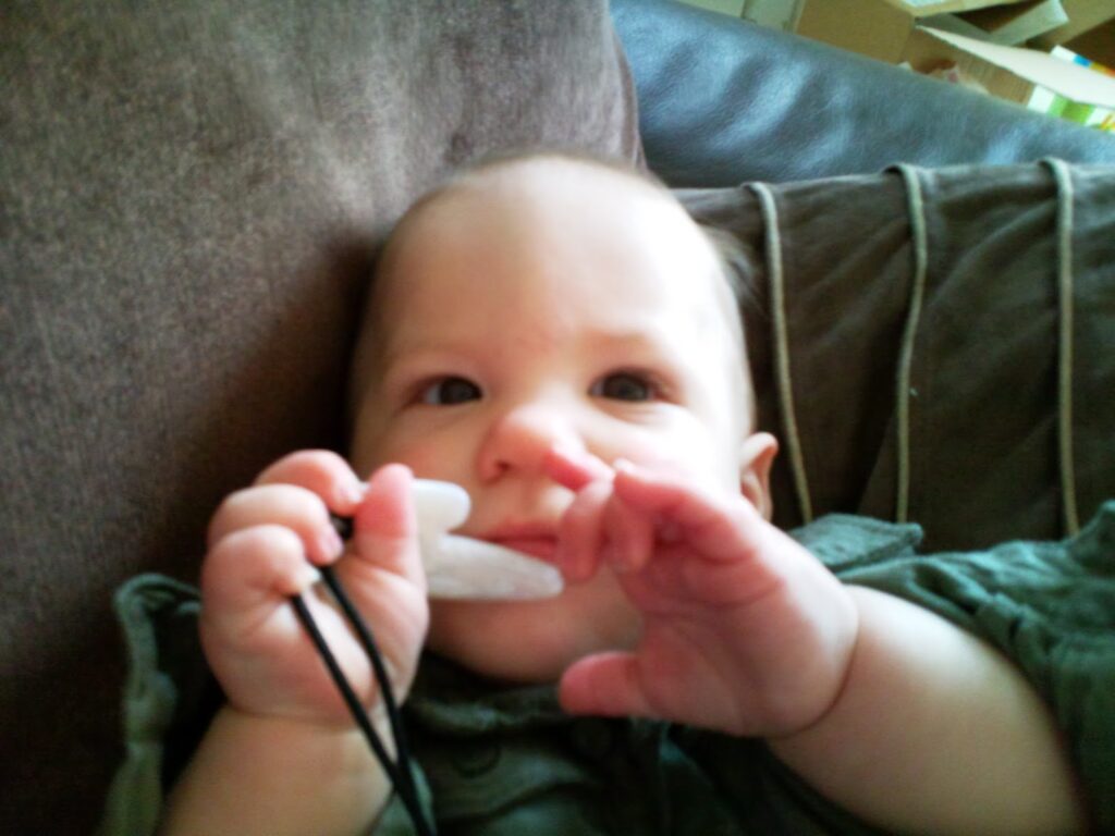 A baby with light skin and blue eyes lies on a brown sofa, holding a white teething necklace with both hands. The baby looks curiously towards the camera, wearing a green outfit. Cushions and some boxes are visible in the background.