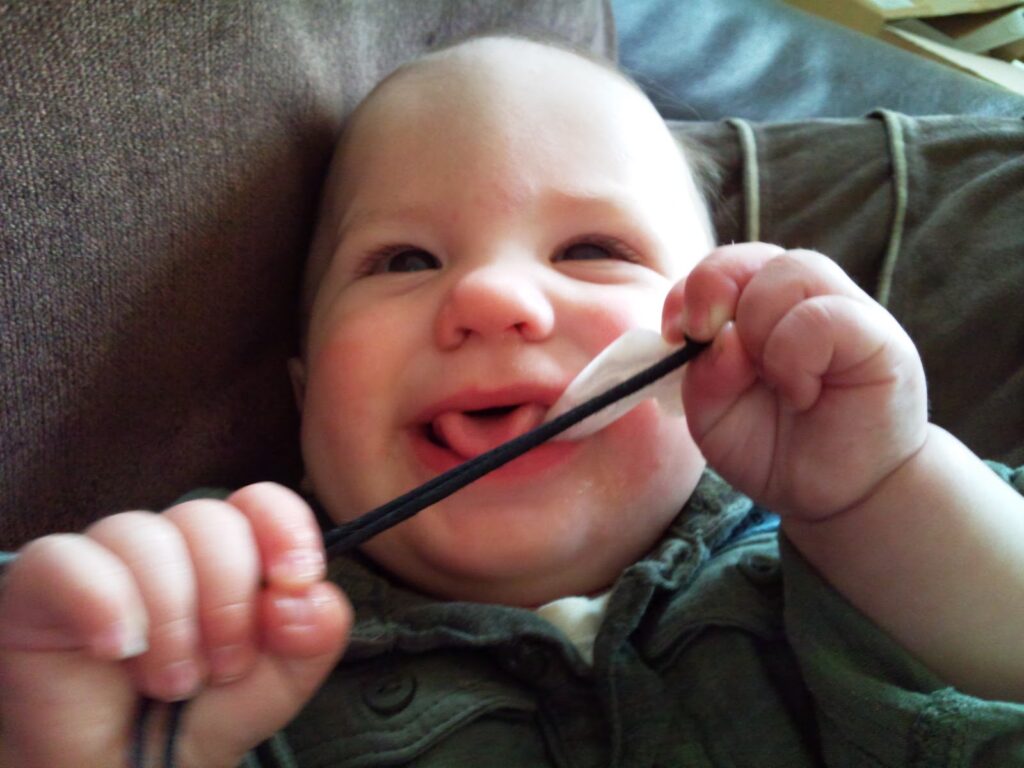 A smiling baby sits on a sofa, holding and chewing on a black lead with both hands. The baby has light skin and is wearing a green jacket. The background is a brown cushion, and the baby appears content and playful.