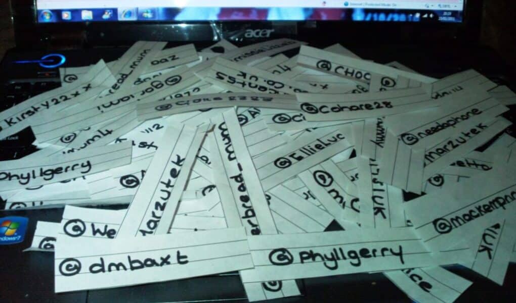 Strips of white paper with handwritten Twitter handles are scattered across a laptop keyboard. Each strip has a different username written in black ink. The laptop screen in the background is partially visible, displaying a mostly white webpage.