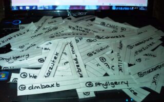 Strips of white paper with handwritten Twitter handles are scattered across a laptop keyboard. Each strip has a different username written in black ink. The laptop screen in the background is partially visible, displaying a mostly white webpage.