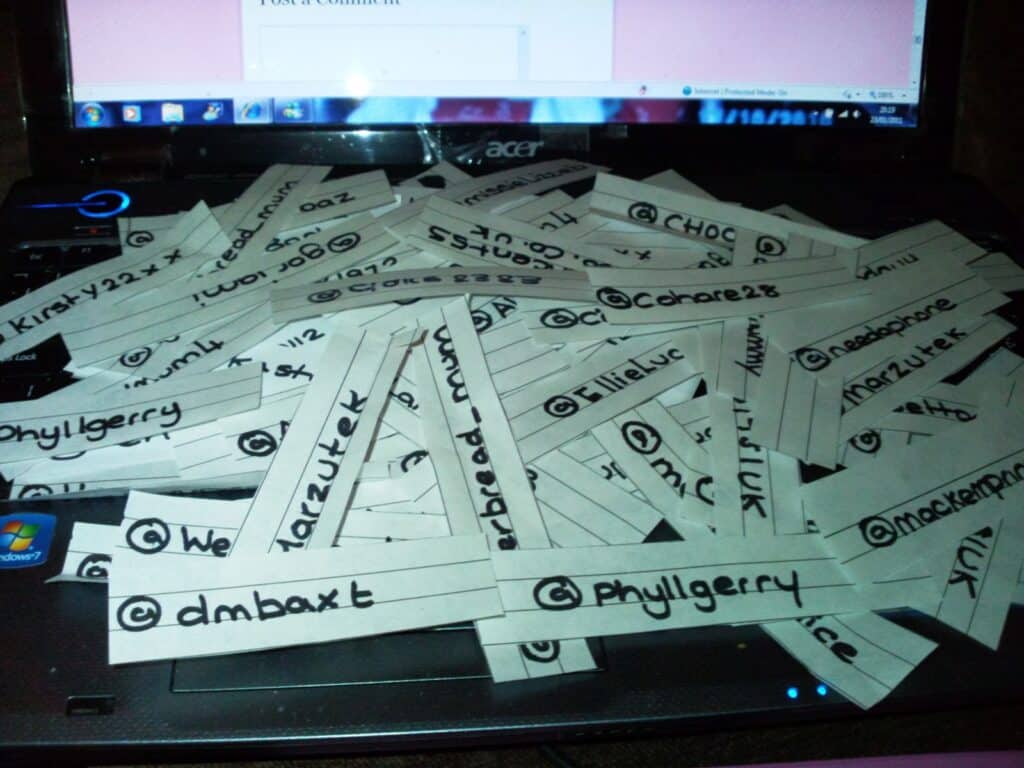 Numerous handwritten strips of paper with various Twitter handles are scattered across the keyboard of an Acer laptop. The laptop screen is partially visible in the background, displaying a mostly pink and white webpage.