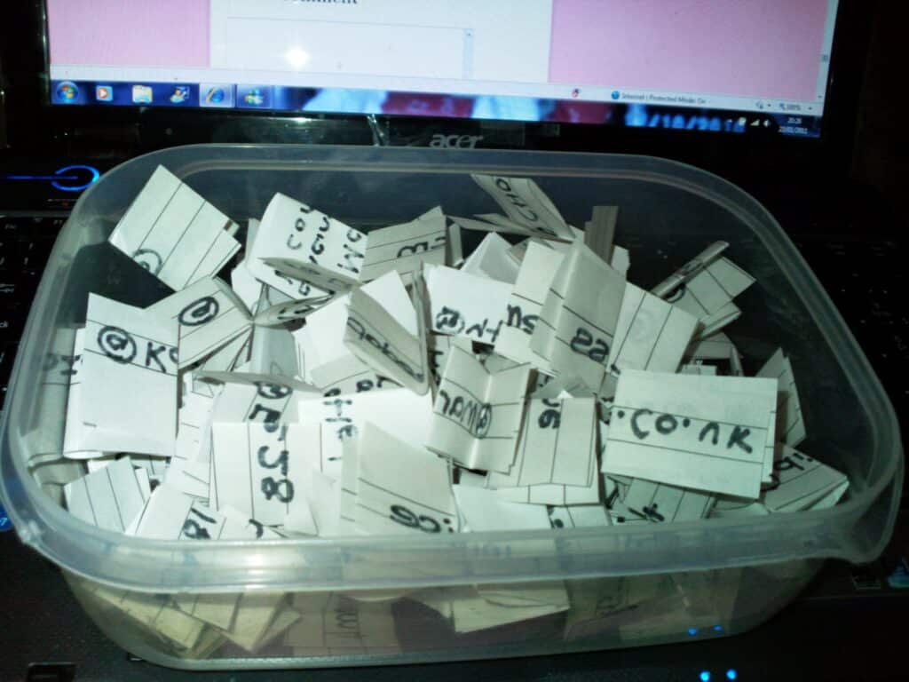 A clear plastic container filled with many small folded slips of white paper, each with handwritten black text or codes. The container is placed on a laptop keyboard, and the laptop screen in the background displays a mostly pink window.