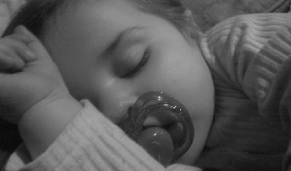 A close-up, black-and-white photo of a young child sleeping, wearing a striped shirt. The child is lying on their side, arm bent near their face, and has a dummy in their mouth. Their eyes are closed and their expression is peaceful.