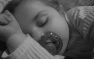 A close-up, black-and-white photo of a young child sleeping, wearing a striped shirt. The child is lying on their side, arm bent near their face, and has a dummy in their mouth. Their eyes are closed and their expression is peaceful.